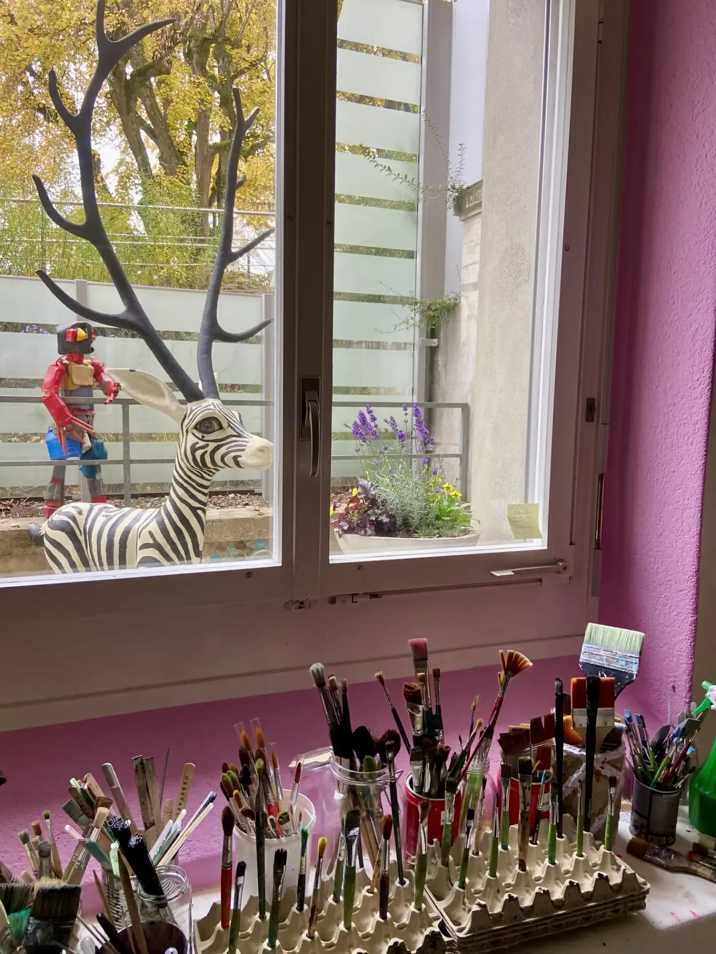 Des dizaines de pinceaux rangés dans des pots devant une fenêtre, un mur rose vif, et dehors une sculpture de zèbre à bois de cerf.